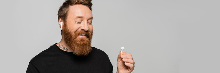 Obraz premium joyful bearded man in black t-shirt listening music and looking at wireless earphone isolated on grey, banner.