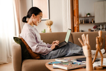 Asian woman working on laptop while laying on sofa in living room