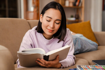 Smiling asian woman reading book while laying on couch