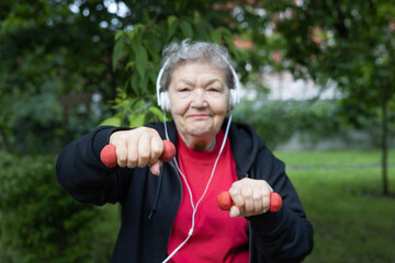 Smiling modern elderly woman in headphones is doing exercises with dumbbells outdoors in the park.  Focus is on hands. Active life of pensioners. Adaptation of pensioners in the modern world. 