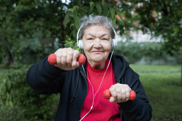Smiling modern elderly woman in headphones is doing exercises with dumbbells outdoors in the park.  Active life of pensioners. Adaptation of pensioners in the modern world. 