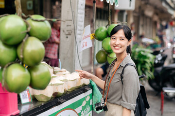 Fototapeta premium Happy young Asian woman backpack traveler stand in front of coconut juice shop at China town street food market in Bangkok, Thailand. Traveler checking out side streets. 