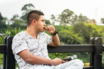 young Latino man, pensive, looking towards the horizon, sitting on a chair in an open-air park.