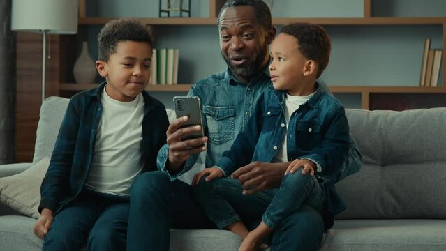 African American family adult father with two school kids children boys kids sons on sofa use funny smartphone apps amazed having fun with technology together look at phone screen video call at home