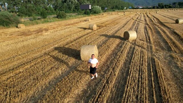 Happy farmer running with hands wide open on agriculture field with round straw bale, aerial shot