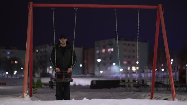 Mentally ill depressed man on empty children playground with swing on iron chains at winter night. Concept of child kids abduction