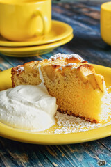 A slice of homemade apple pie with powdered sugar and whipped cream. Served on a yellow plate on a blue wooden table.