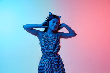 Little beautiful girl, child listening to music in headphones over gradient blue pink studio background in neon light. Concept of childhood, emotions, fun, fashion, lifestyle, facial expression