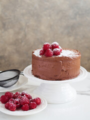 chocolate cake decorated with fresh raspberries and icing sugar. Placed on the table on a white cake stand next to a plate with raspberries and a sieve with powdered sugar.