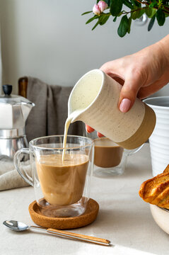 Spring cozy breakfast concept. Two glasses with coffee, croissants, cream is pouring to the glass, woman hand holding pitcher, spring flowers, coffee pot