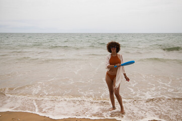 A beautiful adult woman plays tennis on the beach with a blue racket. The woman is on the shore of the beach doing different positions with the racket. Summer sports concept.