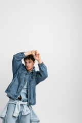 stylish young man in denim jacket and jeans posing with hands above head isolated on grey.