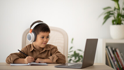 Cheerful european small kid in headphones has video call with teacher, tutor at table in living room