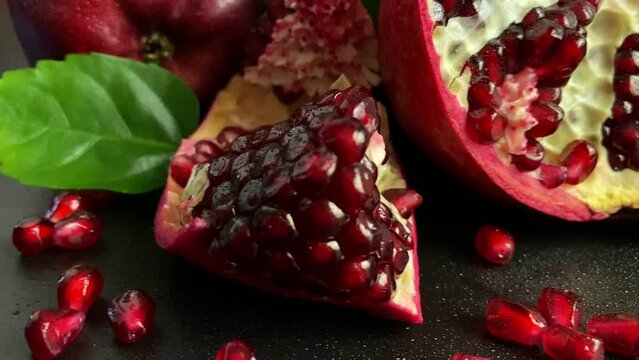 Composition ripe pomegranates, red apples, green leaves on black background. Close-up fresh pomegranate arils. Cut open fruit scattered red grains seed indoors. Studio moving shot. Health nutrition