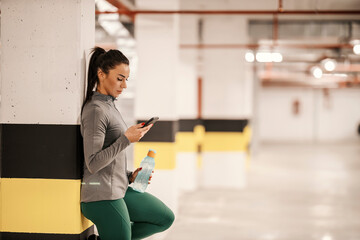 A slim sportswoman in shape is reading messages on the phone while resting from exercises in garage.