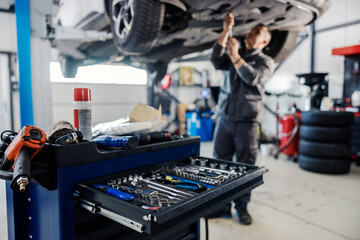 Selective focus on mechanic's toolbox with tools in drawer with auto-mechanic fixing car in blurry background at workshop. - obrazy, fototapety, plakaty