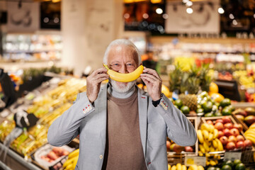 A silly old man is pretending banana is his mouth while looking at the camera. - obrazy, fototapety, plakaty