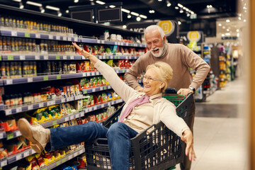 A funny old couple is driving in shopping cart at the supermarket. - obrazy, fototapety, plakaty