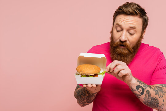 bearded man taking french fries from carton pack with tasty burger isolated on pink.