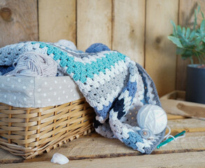 Fototapeta na wymiar white, grey, blue granny square blanket with woolen balls in an white textile basket and crochet hooks on wooden ground
