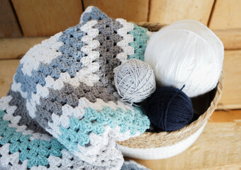 white, grey, blue granny square blanket with woolen balls in an white textile basket on wooden ground