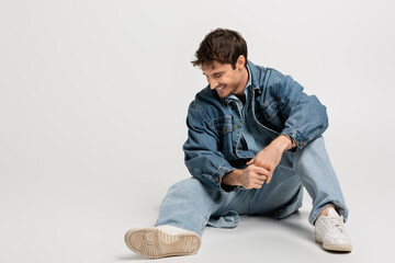 full length of stylish man in denim jacket and jeans sitting and laughing on grey.