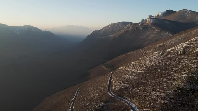 Aerial view of a scenic forest road on mountainside in wintertime at sunset with snow on Mount Terminio, Serino, Avellino, Irpinia, Campania, Italy.
