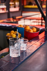 a glass shaker with ice and a bar spoon, a glass for a summer cocktail with ice stands on a blue bar counter against the background of a summer bar with fruits, oranges, lemons, an orange umbrella on 