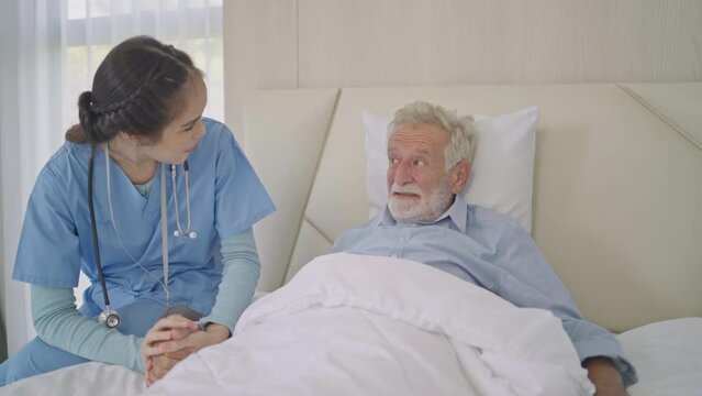 Female caregiver holding hand giving support and empathy to smiling senior man on bed. Happy grandfather relaxing and preparing to sleep