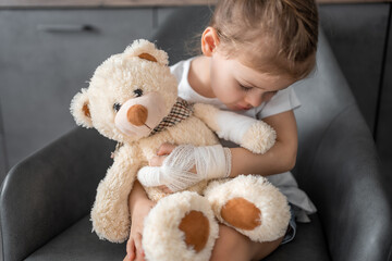 Little girl with broken finger holds teddy bear with a bandaged paw at the doctor's appointment in the hospital