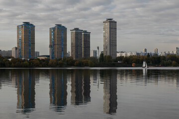 Naklejka premium Landscape of the city lake. Strogino beach, Moscow. A city park along the embankment, several tall apartment buildings are reflected in the water surface. A lone sailboat goes down the river