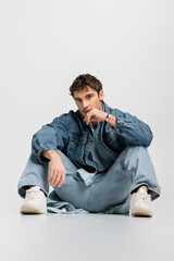 full length of stylish man in denim jacket and jeans sitting and looking at camera on grey.