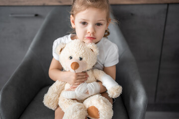 Little girl with broken finger holds teddy bear with a bandaged paw at the doctor's appointment in the hospital