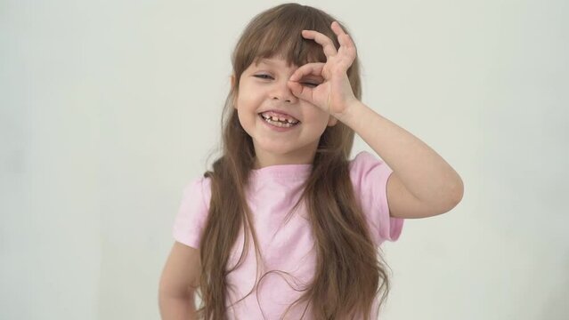 Portrait of a funny little girl with brown hair and a toothless smile, laughing happily and showing OK gesture at her eye isolated on white background. Child girl making Okay fingers. Copy space