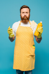 displeased man in yellow apron puffing cheeks while standing with detergent and spray bottle isolated on blue.