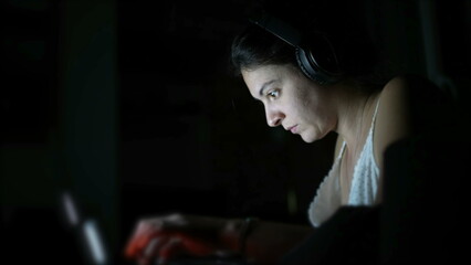 Fototapeta na wymiar Focused woman typing on laptop keyboard in the dark wearing headphones. Person working in the dark. Modern lifestyle