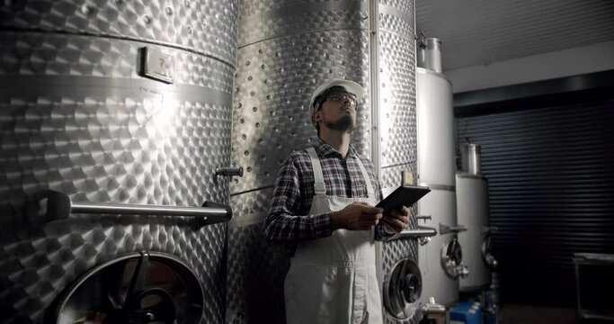 Male wine maker checking quality of beverage distillation process indoors and write down the dates in tablet gadget. Alcohol production concept. Wine production plant. Steel wine distilling tanks.