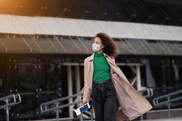 Young african woman girl in wearing face mask ffp2 walking outdoors carrying a suitcase and going...