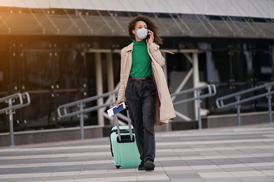 Young african woman girl in wearing face mask ffp2 walking outdoors carrying a suitcase and going to travel by airplane at modern airport. Vacations, travel and active lifestyle concept  