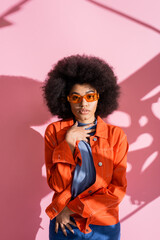 curly african american woman in stylish orange sunglasses and trendy outfit looking at camera on pink.