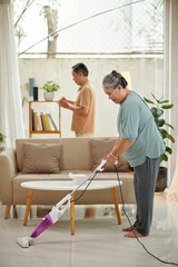 Fototapeta na wymiar Senior woman vacuum cleaning carpet when her husband wiping dust from shelves