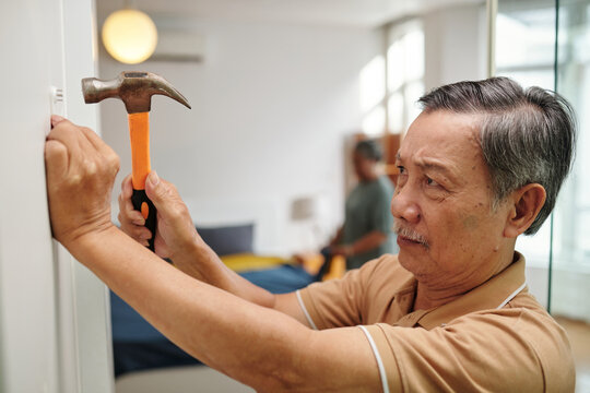 Senior man hammering in nails to hang picture on wall in apartment