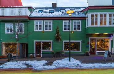 Typical Icelandic green wooden house with snow on the roof in a Reykjavik street and shops with windows with a snow covered sidewalk - obrazy, fototapety, plakaty