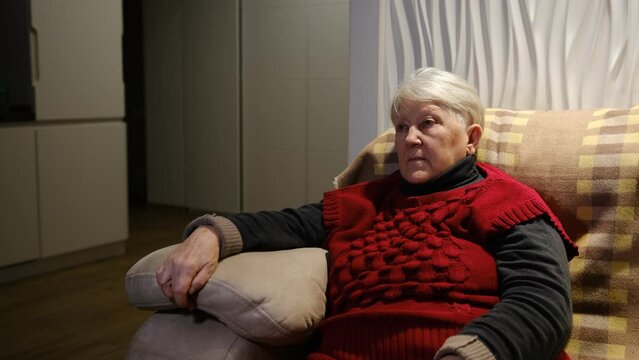 Close-up Of serious Grandmother Watching News On Television At Home. Senior woman in red clothes sitting on biege couch and watch TV