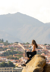 Fototapeta na wymiar beautiful woman sitting and standing on a huge rock with the city or town in the background dressed in black pants and white blouse