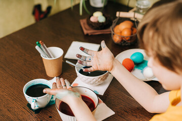 Easter day. Cropped close up Kid boy with dirty hands painting eggs on wooden background. Child sitting in a kitchen. Preparing for Easter, creative homemade decoration.