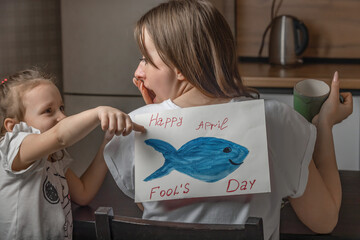A cheerful little girl glues a paper fish to her mother's back at home in the kitchen, a young woman has breakfast, in her hands is a mug of tea. Funny family jokes, April Fool's Day celebration