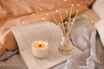 Burning scented candle with glass bottle with home liquid perfume on paper book in bed closeup over glowing lights in bedroom. Aromatherapy. Good morning. - obrazy, fototapety, plakaty