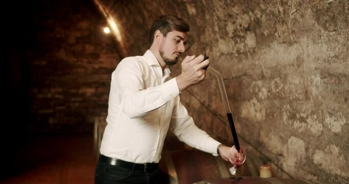 Man testing wine in a factory or warehouse. Winemaking concept. Nicely dressed Sommelier checking on wine by sampling pipette. Sommelier taste expensive drink.