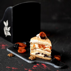 Piece of cake with dried fruits. Dessert with dried apricots and prunes on wooden board. Festive baking. Biscuit cake. Copy space. Side view. Soft focus, dark background.